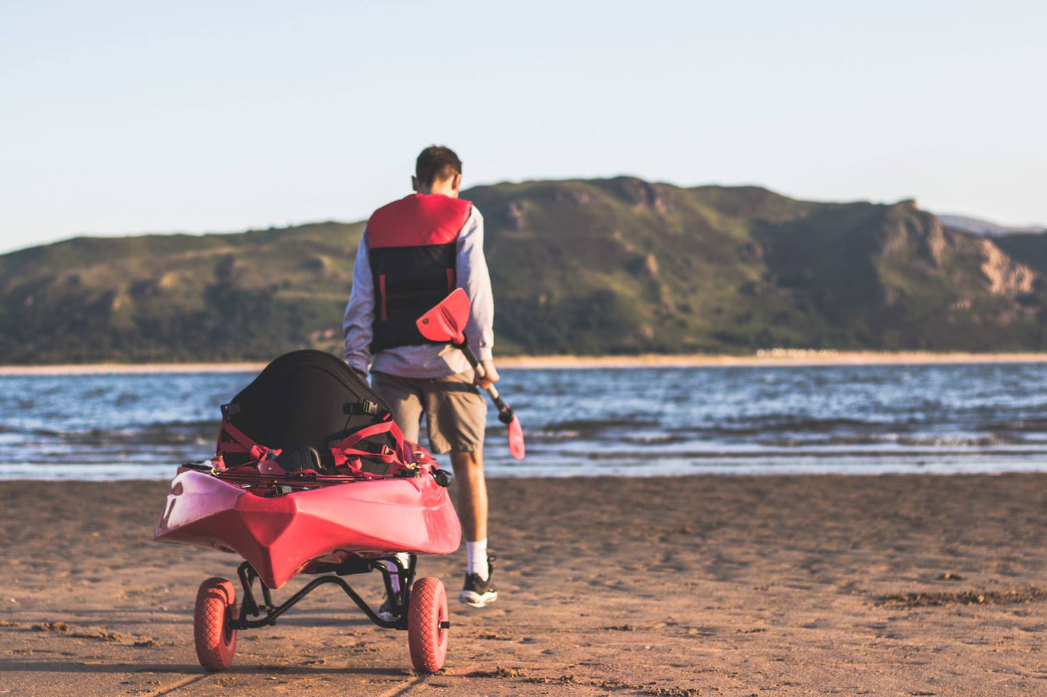 man with a paddle in his right hand is pulling a kayak on trolley down the beach towards the waters edge, there are rocky hills in the background across the water